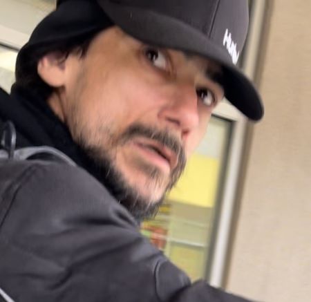 Photo du suspect regardant par-dessus son &eacute;paule droite. Il a des poils au visage et porte une veste noire et une casquette de baseball. 