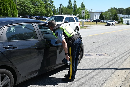 Un policier en uniforme se penche &agrave; la fen&ecirc;tre d&rsquo;une voiture immobilis&eacute;e pr&egrave;s d&rsquo;une &eacute;cole par une journ&eacute;e ensoleill&eacute;e. 