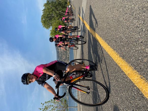 A team of cyclists in black and pink uniforms ride together on a roadway with mountains in the background.