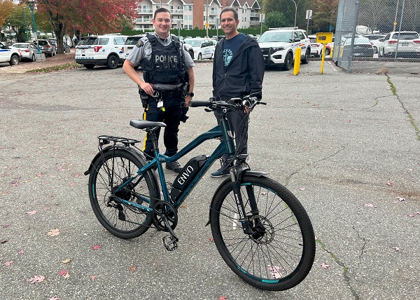 Constable Shaw and Ahmad standing behind the returned blue electric bicycle with marked RCMP vehicle&rsquo;s in the background