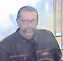 A Caucasian man with slicked back brown hair and a full beard, wearing glasses and a black jacket with a red and white horizontal stripe on the front.