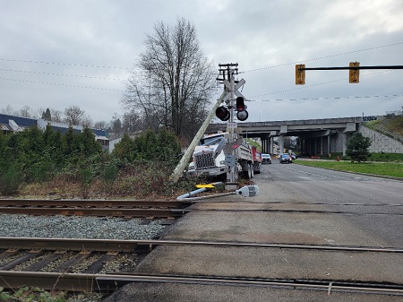 A front view of a dump truck that had just collided with a hydro pole and a lamp post on the side of the road, just before a train track and the SkyTrain. The photo is taken from the other side of the train track.