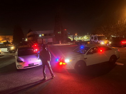 Man standing in front of a white vehicle which is next to a white police car with its emergency lights flashing