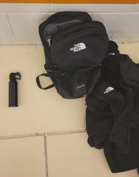 A black canister of bear spray, a black bag and a black jacket on a yellow tile floor.