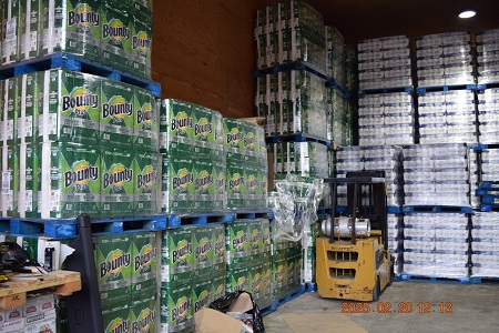 Skids of paper towel and toilet paper stacked in a warehouse with a yellow forklift parked in front of it.
