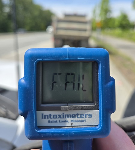 A blue roadside screening device with the word FAIL on it with a blurry dump truck in the background, parked on the side of the road.
