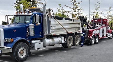 A dump truck with a blue cab is hooked up to a red tow truck in a parking lot.