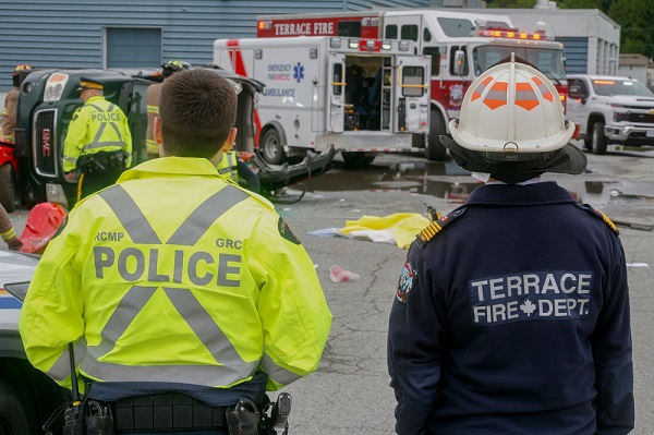 A police officer and firefighter look over a mock collision scene with twisted cars and student actors