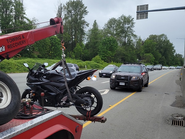 A black motorcycle is towed to the impound yard as a BC Highway Patrol cruiser is visible in the background