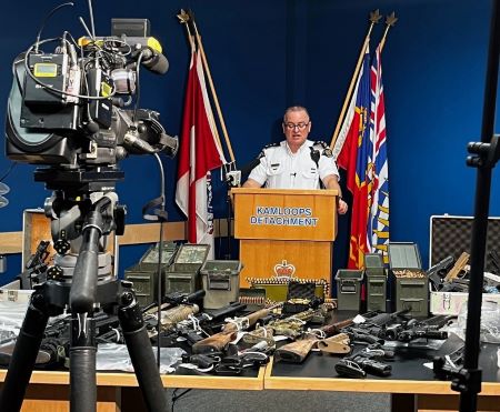 Supt. Pelley stands behind a Kamloops Detachment podium while speaking to the local media. Items seized from a recent investigation are displayed on two tables in front of him. 