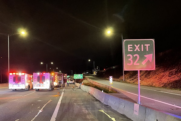 Photo of two ambulances and a firetruck with lights on at night time blocking two lanes of the highway ahead of the sign for <q>Exit 32</q>.