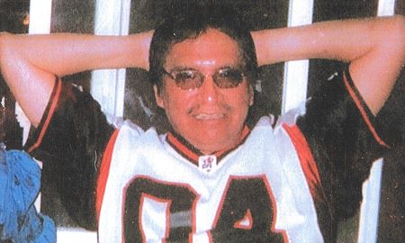 Fred Yellow Old Woman est assis, les mains derri&egrave;re la t&ecirc;te. Il porte des lunettes fonc&eacute;es et un chandail de sport noir, blanc et rouge avec le num&eacute;ro &laquo; 04 &raquo; sur le devant.