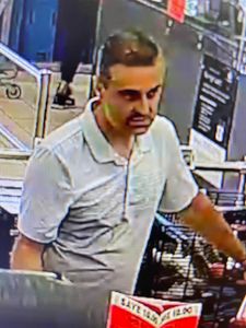 male suspect inside the store. He is wearing a light coloured gold shirt. He has a buzz cut with big dark eyebrows, dark coloured hair and 5 o&rsquo;clock shadow on his face.