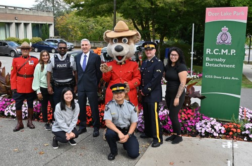 A group of people, including several RCMP officers in uniform smile while standing outdoors with a bear mascot wearing a Red Serge beside a sign for the Burnaby RCMP detachment