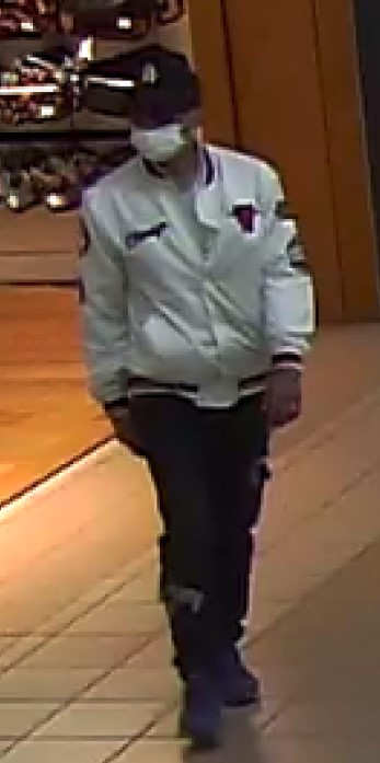 A man facing the camera, wearing a dark LA Dodgers baseball hat, white medical mask, white bomber jacket with a Chicago Bulls crest, black pants and dark shoes.