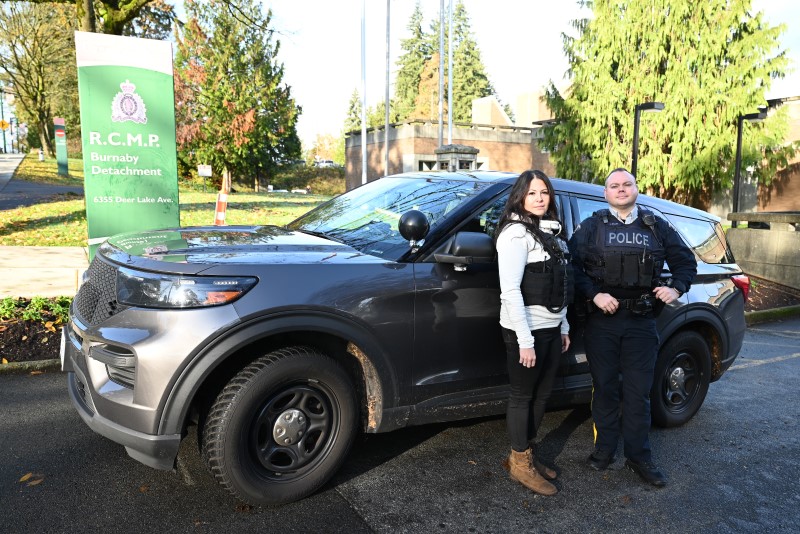 A man and woman wearing vests stand in front of a police SUV on a sunny day
