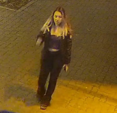 : Photo d&rsquo;une femme qui accompagnait l&rsquo;homme. Dans cette image, ses cheveux longs sont d&eacute;tach&eacute;s et son manteau fonc&eacute; est ouvert. Elle porte un chandail court et des pantalons fonc&eacute;s. Son manteau a un logo blanc en &eacute;cusson coll&eacute; sur son &eacute;paule gauche. 