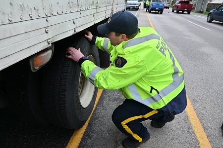 A police officer wearing a dark blue baseball cap and a yellow traffic jacket inspects the tire of a commercial vehicle.