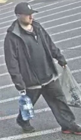 Caucasian man walking through a parking lot, wearing a black baseball cap, dark sweater, dark jacket, jeans and black shoes.