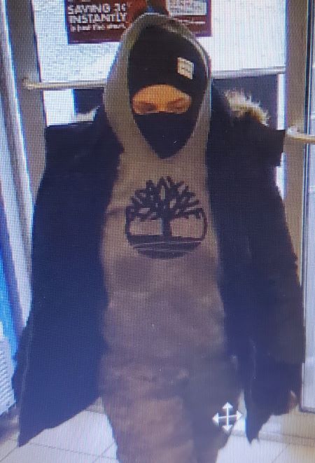 A woman with a mask, dark jacket, and a greyish sweater with a black tree and circle logo. 